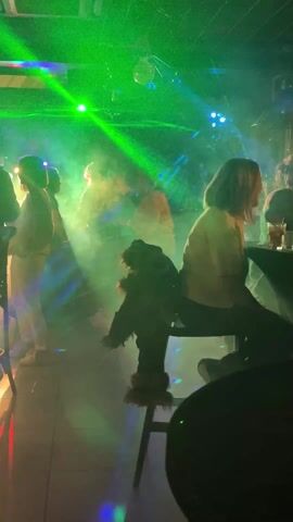 The video depicts a dimly lit bar with several people seated at tables. Disco lights, including green laser beams, are shining throughout the space. The first shot shows a few individuals standing or sitting near a table, with a furry puppet-like object propped up on a chair. The next sequence pans to other tables occupied by groups of people, engaged in conversation. One man is prominently featured, seated at a table with three partially filled glasses of colored liquid. The lighting shifts, casting a green hue on the man.