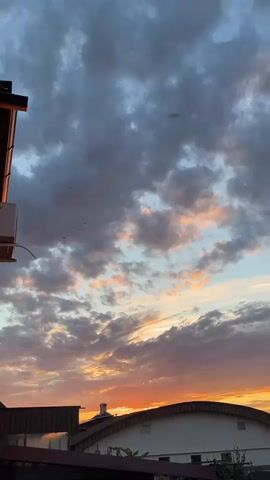The video shows a panoramic view of a cloudy sky at sunset. The clouds are illuminated by the setting sun, casting hues of orange, pink, and purple across the sky. Below the sky, there are buildings with roofs and some trees. There are no people visible in the video. The camera is static and the video captures a slow pan from left to right.