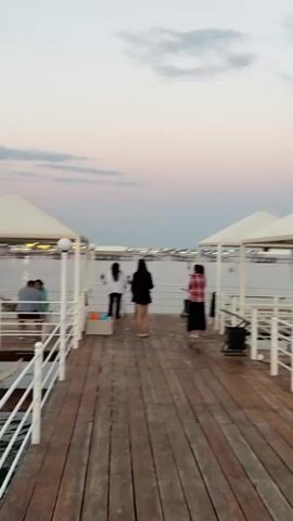 A group of people are gathered on a wooden pier over water. The sky is a soft gradient of pink and grey, suggesting either sunrise or sunset. On the left side of the pier, under a white canopy, two people sit at a table. Further down the pier, a group of four individuals stand near the railing, one of whom is wearing a black outfit and the other a white outfit. To the right of this group, another person in a red and black plaid shirt stands near a black trash can. In the foreground, the pier is made of weathered wooden planks. The water is calm, and structures, possibly buildings or other piers, are visible in the distance across the water, with lights illuminating them. The overall atmosphere appears relaxed and social.