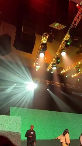 The video shows a stage with professional lighting and sound equipment. There are two people on stage, a man and a woman. The man is standing on the left, facing the audience and appears to be speaking. The woman is standing on the right, facing away from the audience and gesturing with her hand. They are both dressed in casual attire. The stage is decorated with a textured green backdrop, suggesting an event or performance is taking place. The lighting is dramatic, with spotlights creating beams of light across the stage. The overall impression is of a dynamic and engaging event.