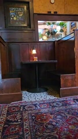 The video shows the interior of a pub or restaurant featuring wooden booth seating. The camera slowly pans right, showing multiple booths arranged in a row. Each booth is made of dark wood and includes a small table with a black base and a single wall-mounted lamp. The floor is covered with small white and black tiles, and a colorful, patterned rug lies in the foreground. The rug features a mix of red, blue, and brown colors. A small window separates the booths, allowing a view of plants in the background. In the distance, a bar with bottles and glasses can be seen. There are no people visible in the video.