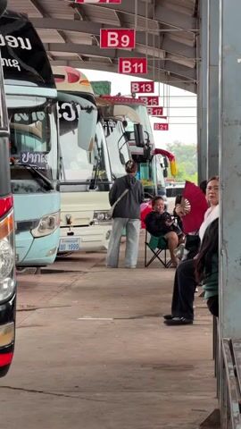 A bus station scene is depicted. Several buses are parked in a covered bay. Red signs with white lettering indicate bus bay numbers, such as B9, B11, B13, B15, and B21. In the foreground, on the right, a person is seated on a bench, partially obscured by a pillar. Their head is turned towards the left, and they are wearing dark clothing. To the left of this person, another individual is seated in a folding chair, holding a smartphone and a fan. They appear to be looking at the phone. Further to the left, a person in a dark jacket and light-colored pants is walking away from the camera. They are carrying a green plastic bag. The ground appears to be concrete. The lighting suggests an overcast or shaded environment.