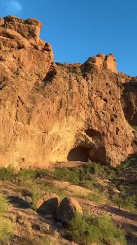 The video shows a rocky desert landscape with sparse vegetation under a clear blue sky. A large, reddish-brown rock formation dominates the left side of the frame, featuring a cave-like opening. The ground is uneven, with loose rocks and dry brush. The lighting suggests it is either early morning or late afternoon, casting long shadows. The camera pans slowly to the right, revealing more of the rugged terrain and distant hills. No people or animals are visible in the video.