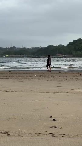 A lone figure walks along a sandy beach near the water's edge. The person is silhouetted against the ocean, which has moderate waves. In the background, a lush green hillside rises, dotted with buildings and trees. The sky is overcast and gray. The person appears to be carrying a long, thin object, possibly a fishing rod, horizontally across their body. The sand in the foreground is textured with ripples and scattered debris.