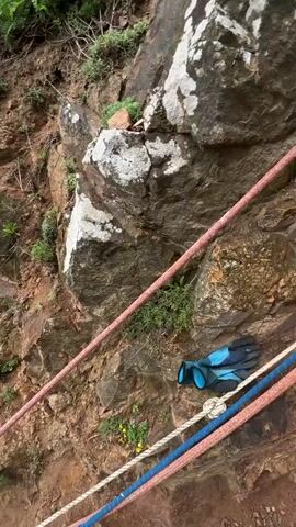 The video shows a woman standing on a rocky slope looking towards a small beach. The slope is steep and there are ropes running down the path for support. The beach has red sand and is surrounded by rocky cliffs. The sea is visible in the distance. The woman has long dark hair and is wearing a white top and a black fanny pack.