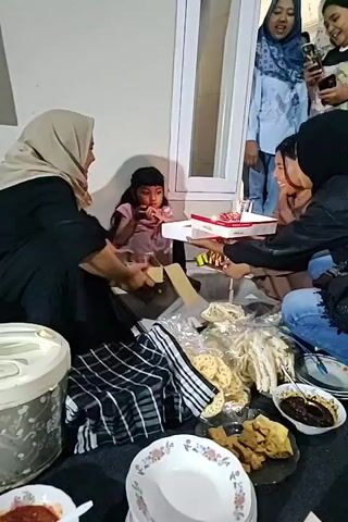 A video shows a group of people gathered for a birthday celebration. A young girl wearing a pink shirt is sitting on the floor, holding a birthday cake with lit candles. Two women are sitting next to her, one on her left wearing a beige hijab and black clothing, and another on her right wearing a black hijab and a denim jacket. They are both looking at the girl and the cake. Behind them, standing up, are three other women. One woman in a blue hijab and light blue top is holding a phone and looking towards the girl. Another woman next to her, wearing a black top, is also looking at the girl and holding a phone. The third woman is slightly out of focus and appears to be smiling. In the foreground, on the floor, there are plates with food, including crackers and what appears to be fried food, as well as a large plastic container on the left. The gathering appears to be taking place indoors, with a neutral-colored wall and a window visible in the background. The lighting suggests an evening or indoor setting. The overall atmosphere seems celebratory and intimate.