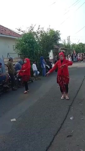 The video shows two women in red outfits dancing in the middle of a paved road. They are wearing headscarves and what appear to be traditional skirts. The woman on the right is facing the camera and her arms are outstretched. The woman on the left is also dancing and her arms are in a similar position. There are people watching the dancers from the side of the road, some sitting on motorcycles. There are also houses and trees visible in the background. The scene appears to be outdoors during the daytime.
