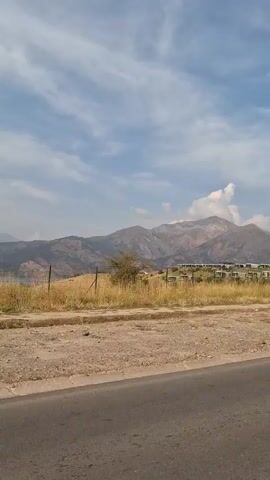 A black car is seen driving on a road. In the background, there is a lake, mountains, and a paraglider in the sky. The sky is partly cloudy.