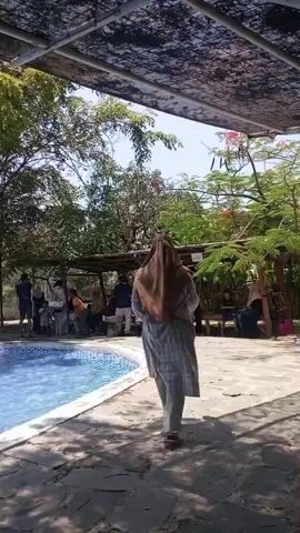 A group of people are gathered near a swimming pool. A woman wearing a headscarf walks away from the camera, towards a covered seating area. Several people are sitting and standing in the shade of the covered area. The swimming pool is to the left of the frame. Trees and foliage surround the area, and the sky is visible.