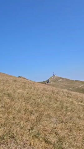 The video shows a panoramic view of a mountain landscape under a clear blue sky. In the foreground, dry, tall grass covers rolling hills. A dirt path winds through the grass. In the distance, more rolling hills are visible, some covered in green vegetation and others with dry grass. Further back, a range of dark green, forested mountains stretches across the horizon, with lighter blue, hazy mountains behind them. A few people can be seen walking on the path in the middle distance. On the summit of one of the distant peaks, there are some structures, possibly radio towers or a weather station.