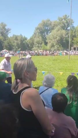 This is a medium-shot, outdoor video taken on a sunny day with a clear blue sky. The scene appears to be a festival or outdoor event. In the foreground, several people are seated, facing away from the camera, presumably watching an event unfold on a grassy field. They are dressed casually, with some wearing hats and headscarves. The middle ground of the video shows a lively scene. A woman wearing a white dress and a headscarf is carrying two buckets on a pole across her shoulders. She is walking past a group of people, also dressed in casual attire. Behind her, a man wearing a white patterned shirt and a cap is walking, and it seems he might be participating in a race or game. A blue flag is flying on a tall pole in the background. Further in the distance, a large crowd of people are gathered, and there are tents set up. The environment is characterized by a large expanse of green grass and numerous trees in the background, indicating a park or rural setting. The overall atmosphere suggests a community gathering or celebration.