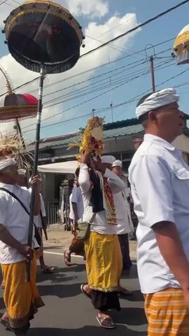 A procession of people walk down a street. In the foreground, a man in a white shirt and orange sarong carries a tall pole with a large umbrella on top. Behind him, others in similar attire carry offerings and decorations, including ornate parasols and tall, curved wooden structures adorned with smaller decorative elements. Several people are wearing white shirts and sarongs, while some women wear traditional Balinese white blouses with sarongs. The procession appears to be part of a cultural or religious event, possibly a parade or ceremony. The environment is a street with buildings and power lines visible in the background, suggesting an urban or semi-urban setting. The weather is sunny with some clouds.