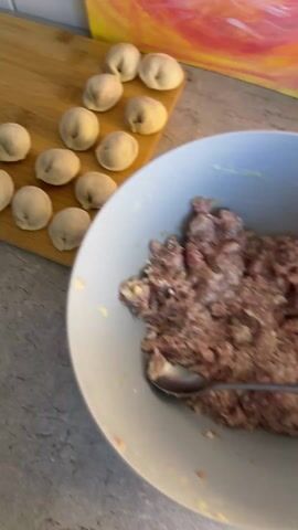 The video shows someone preparing what appears to be Russian pelmeni or dumplings. The video begins with a close-up of a wooden cutting board with several finished pelmeni on it, along with a plate of meat filling. The camera then moves to a countertop dusted with flour, where several rounds of dough have already been cut. A person wearing a white long-sleeved shirt picks up a round of dough and begins pinching it together, forming a half-moon shape. Other kitchen items, such as a rolling pin and a covered mass of dough, are visible in the background. The kitchen has white tile and a gray countertop, and a colorful item, possibly packaging, sits behind the cutting board at the start of the video.