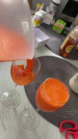 A close-up shot shows a clear pitcher pouring orange juice into a clear wine glass. The glass is one of two on a grey woven placemat on a white table. Pills, a green box, a bottle of honey, and red scissors are also visible on the table. The juice has a foamy appearance. The pitcher is being held offscreen.