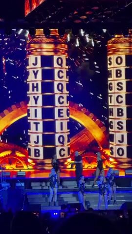 A video opens with a large, bright, fiery graphic on a screen behind a stage. On the stage, there are several people, including a man singing and a group of women dancing in costumes. The video then transitions to show two large, rotating columns made of blocks with letters on them. Confetti falls from above. The man continues to sing, and the dancers continue to perform. The stage is lit with bright blue and white lights.