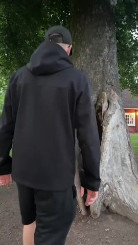 A person in a black hooded jacket and black shorts is climbing into a hollowed-out tree trunk. The person's back is to the camera, and they are stepping into the opening with their right leg. The tree is large and has a rough, weathered exterior. The ground around the tree is dirt and grass. In the background, there is a red building with white trim and a green tree line. The scene appears to be outdoors during the daytime.