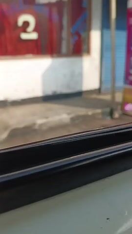 THE VIDEO BEGINS WITH A BLURRED VIEW OF A BLUE SHUTTERED DOOR WITH A LARGE WHITE X AND PINK SIDE PANELS. THE SCENE THEN TRANSITIONS TO A VIEW THROUGH A CAR WINDOW, SHOWING A FENCE AND BUILDINGS OUTSIDE. FLAGS ARE VISIBLE IN THE BACKGROUND. THE VIDEO CONTINUES TO SHOW A STREET SCENE WITH MOTORCYCLES, BUILDINGS, AND PEDESTRIANS. THE CAMERA IS POSITIONED INSIDE A VEHICLE, LOOKING OUT A WINDOW.