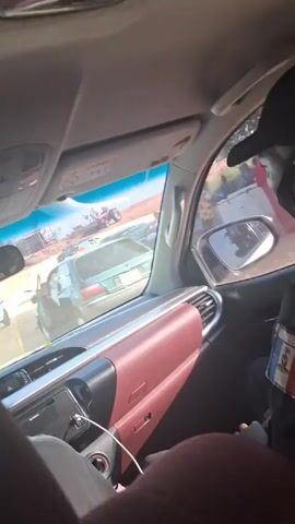 The video shows the inside of a vehicle, likely a car or truck, with a maroon dashboard. Through the windshield and side window, an accident scene is visible. People are standing around two damaged vehicles. The first is a white van with damage to its rear end. The other vehicle is partially visible and appears to be a pickup truck. The accident scene is on a dirt road in what appears to be a village or small town. There is also a motorcycle parked nearby. Inside the car, the passenger has an official-looking armband and can be seen holding something. There is also a small air freshener hanging from the rearview mirror with a person's picture on it.