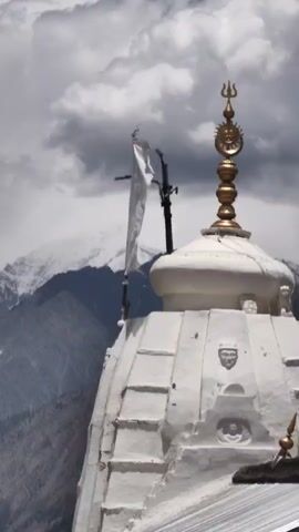 The video shows a close-up view of the top of a white temple structure, likely in a mountainous region. The temple has a conical roof with decorative elements, including what appears to be a trident at the very top, along with a golden sun symbol. A white flag is attached to a pole near the temple's peak, fluttering in the wind. Behind the temple, a snow-capped mountain range is visible under a cloudy sky. The lighting suggests it is daytime. The focus is on the temple and its surroundings, with no people or animals visible in the frame.