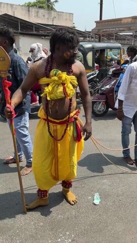 The video depicts a street scene in India, with several people gathered around. A man in traditional attire, wearing a yellow dhoti and adorned with beads and turmeric, stands in the foreground, holding a wooden staff. Next to him, a man in a white shirt and jeans stands barefoot. In the center of the scene, a woman in a pink salwar kameez picks up a black plastic stool from the ground. Behind her, a small altar-like structure decorated with coconuts and peacock feathers is visible. A young boy stands beside the woman, looking at the altar. The background shows a busy street with auto-rickshaws and other vehicles. Several other people are present, watching the scene.