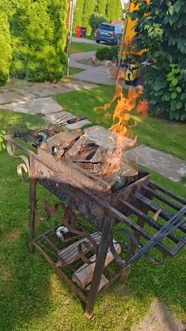 A backyard barbecue grill with burning wood and flames is in the foreground. The grill is made of dark metal and has a grate on top. In the background, a green lawn, a paved walkway, a parked car, and trees are visible.