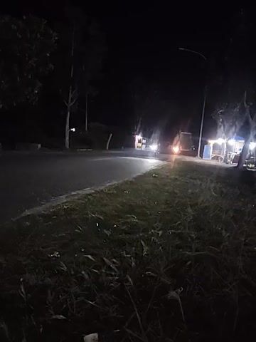 The video is a low-light recording of a nighttime street scene. A paved road curves from left to right, bordered by a grassy verge with tall grass. Trees are visible on the left side of the road, and further down on the right side, there are several stalls with lights illuminating them. A streetlight is also visible. A motorcycle with its headlights on approaches from the distance, crosses the frame from left to right, and then appears to turn or move out of frame. The motorcycle is illuminated by its own headlights, and its motion is blurred due to the low light conditions and possibly movement of the camera. No people are clearly identifiable, though there may be figures at the stalls on the right. The overall atmosphere is dark and somewhat quiet, typical of a rural or less populated area at night.