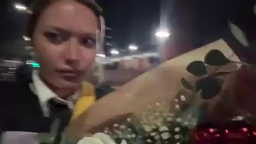 A person is holding a bouquet of flowers and recording a video. The bouquet contains white and red roses, along with baby's breath and eucalyptus. The person is wearing a black jacket over a white collared shirt with a black tie and a yellow sash. They are outside at night, with streetlights visible in the background. There appear to be buildings and some greenery visible as well. The person smiles at the camera.