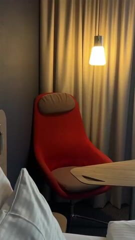 The video provides a tour of a modern hotel room. It opens on a vibrant red armchair with a light brown headrest and seat cushion, positioned against a grey wall. A small, light-wood table with a semi-circular top is next to the chair. Above, a minimalist hanging light fixture emits a warm glow, illuminating the light grey curtains behind the chair.

The camera then pans right to reveal a window covered by sheer white curtains, through which diffused daylight is visible. Continuing the pan, a wall-mounted flat-screen television comes into view, displaying "WDR 1LIVE DIGGI" with "WDR" also visible in the upper left corner. Below the TV, a white shelf holds a black electric kettle, two white mugs, and various small packets, indicative of a hot beverage station. A black, foldable luggage rack stands below the shelf. The room's decor combines grey walls with light wooden paneling. A corner of a bed, dressed in white bedding with a striped pillow, is visible in the foreground throughout the pan. The scene depicts a typical, clean, and functional hotel room setting.