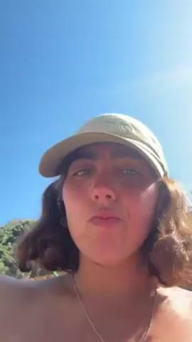 Here's a description of the video:

The video shows a young woman with brown hair and a light green baseball cap, standing outside on a sunny day. The camera is close to her face, capturing her expressions. In the background, there's a building with a light yellow exterior and a red roof. Some greenery is visible behind the building. At one point, a hand reaches into the frame and briefly touches the top of her head. She sticks her tongue out.