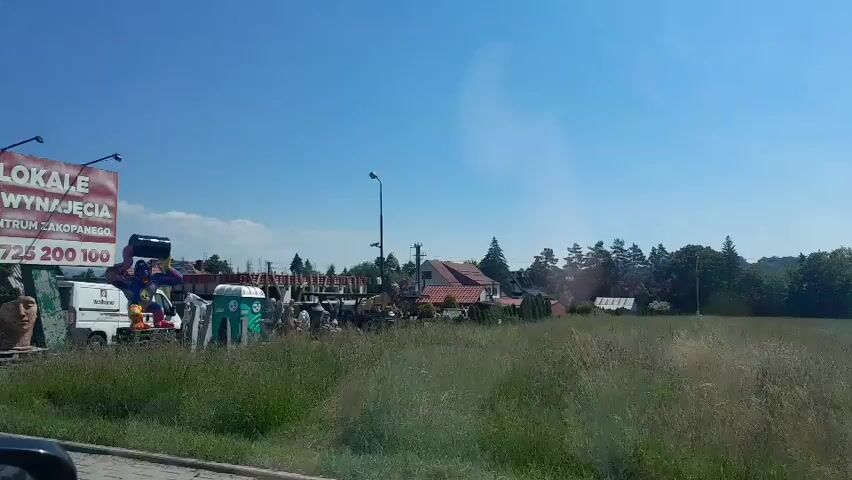 The video shows a roadside scene on a sunny day with a clear blue sky. In the foreground, a grassy field stretches out to the right. A paved surface is visible in the lower left corner. To the left, there is a large billboard advertising "LOKALE DO WYNAJĘCIA W CENTRUM ZAKOPANEGO" (Premises for rent in the center of Zakopane) with a phone number. Beside the billboard, there is a collection of items, including what appear to be garden decorations or souvenirs, such as large statues of animals like a giraffe and a horse, as well as figures of people and a lion. A portable toilet is also visible among these items. Further back, there are several structures, including a green portable building and other makeshift shelters, along with more parked vehicles. A street lamp stands in the middle ground. The overall impression is of a commercial area, possibly a market or a storage yard, situated alongside a road.