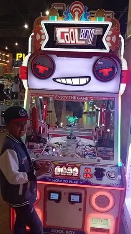 The video shows a child standing in front of a "COOL BOY" claw machine game. The machine is brightly colored with a cartoon robot face at the top. It has a glass enclosure filled with prizes. Below the enclosure is a control panel with buttons and a "HOW TO PLAY" sign. The child smiles at the camera and fiddles with something in his hand. The surrounding environment appears to be an arcade or entertainment center, with other games and people visible in the background.