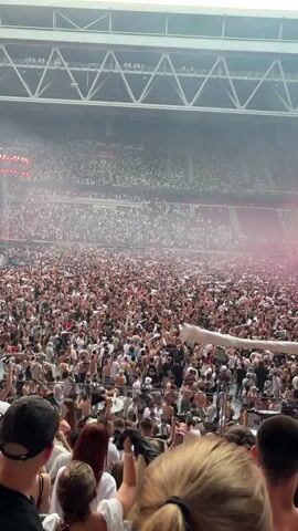 This is a vertical video capturing a large crowd of people at an outdoor stadium event, likely a concert or festival. The scene is filled with thousands of attendees, many of whom are wearing light-colored clothing, contributing to a bright atmosphere. The overall mood appears energetic and excited, with many people raising their hands and some throwing articles of clothing into the air, as seen in the foreground and mid-ground. Smoke or fog, tinged pink by stage lighting, drifts through the crowd.

Prominent features of the setting include the stadium's tiered seating, filled with spectators, and a large stage structure in the distance with numerous lights and sound equipment. The overhead stadium roof structure is also visible, suggesting an indoor or covered portion of the venue. The lighting suggests it is daytime, possibly late afternoon or early evening, given the mix of natural light and stage lighting.

In the foreground, several individuals are clearly visible, looking towards the stage. One person in the lower left is wearing a dark t-shirt and a black baseball cap. To their right, a person with blonde hair is looking forward, and another person's shoulder and head, wearing a black bucket hat adorned with pink flamingos, is prominent in the lower right.

There is no visible text in the video. The event is taking place in Dusseldorf, Germany.