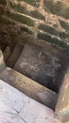 The video pans down a set of worn stone stairs within a stone structure. The walls are made of rough-hewn stones, some of which are covered in green moss and water stains. The steps are also made of stone, with significant wear and discoloration. The light at the bottom suggests an opening or exit. There are no people visible in the video.