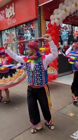 A group of dancers in colorful traditional outfits perform on a sidewalk in front of a red storefront. The dancers, both male and female, wear intricately patterned ponchos and skirts with vibrant embroidery and tassels. Many of them also wear red hats with colorful feathers. They are holding long sticks with fluffy, multi-colored plumes, which they are twirling as they dance. Red and white balloons are strung up above the storefront. The store appears to be a clothing store called "RIBOL". The ground is paved and there are some orange traffic cones visible. The overall atmosphere seems festive.