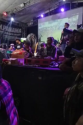Here's a concise description of the video:

The video shows a live performance of a band on a stage. The band consists of several musicians playing instruments, including what appear to be traditional Indonesian instruments. There is a screen behind them displaying a video. Overhead lighting rigs are visible.  The camera appears to be handheld and from a low angle, positioned among the audience members watching the performance. People are standing around the camera's position, observing the band. The stage setup and lighting indicate a professional concert or festival setting.