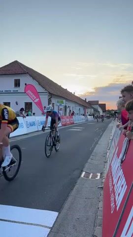 The video shows a bicycle race in progress. Several cyclists are seen riding down a paved road lined with spectators and barriers. The sun is setting, casting a warm glow over the scene. Buildings, including a pink one with a pink teardrop-shaped banner, are visible on the left side of the road. The spectators are standing behind a red barrier on the right side of the road, some are children. The cyclists are wearing racing attire and helmets. The event appears to be well-organized, with clear signage and safety measures in place.