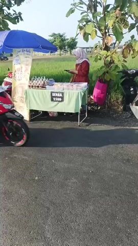 A person wearing a red hijab and a maroon shirt is sitting on a red and black motorcycle, facing left. They are wearing blue pants with white stripes and white sneakers. A white cooler and a clear container with items are strapped to the front of the motorcycle. In the background, another person in a maroon top and a light-colored hijab is standing behind a folding table covered with a green cloth. The table displays numerous small, foil-wrapped food items in rows. A sign on the table reads "SERBA 2500". A blue umbrella is propped over a stand behind the motorcycle. A large poster is attached to the stand. A tree with green leaves is to the right of the table, and a pink bag hangs from its branches. A rice paddy field is visible in the distance. The ground is paved with asphalt.