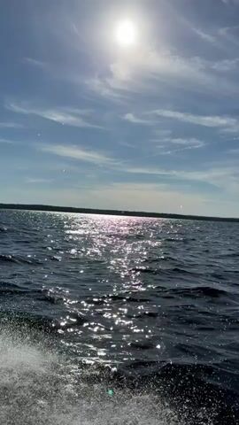 The video shows a view from a boat moving on dark blue water. The sun is bright and high in a blue sky with wispy clouds. The sun's reflection shimmers on the water's surface. A wake of white spray is visible in the foreground, indicating the boat's movement. The horizon is visible in the distance, with a line of trees or land. The scene appears to be a bright, sunny day.
