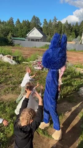 A group of children are gathered in a yard with a furry blue monster costume. One child in a white dress is in the center of the frame, while another child wearing a black jacket is to the left, looking down. A third child with braided hair is in the foreground, also looking down. In the background, a person wearing the blue monster costume stands with their arms raised. Another child in a pink and white dress stands to the right of the monster costume, and a person in a green shirt is visible behind them. The yard appears to be undergoing construction, with dirt piles and wooden structures. A bicycle with a pink frame is parked in the background. The sky is clear with a few clouds, and trees line the perimeter of the yard. The sun is shining, casting shadows across the scene.