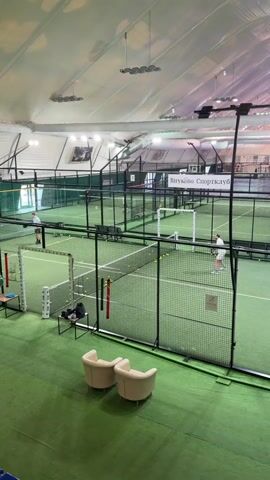 The video shows an indoor Padel tennis court with two people practicing. The court is enclosed with black wire mesh fencing and has a green playing surface. There is a net dividing the court in half. There are tennis balls scattered across the court. Two white chairs are in the foreground. A sign that reads "Vnukovo Sportclub" is visible in the background. Overhead lighting illuminates the scene. One person is standing near the back of the court on the left side, while the other is near the net on the right.