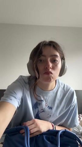 A young woman sits cross-legged on a bed in front of a white wall. She is wearing a light blue t-shirt with a drawing on it, dark pants, and gray headphones. She holds up a blue, gathered leather bag, turning it to show its shape. She then displays a white tag attached to the bag, bringing it close to the camera to show the text printed on it. The bed has a brown and white patterned blanket.