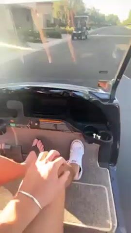 The video shows a male, likely a young adult, driving a golf cart down a paved street with houses and landscaping visible in the background. He is wearing a white t-shirt, sunglasses, and appears to be driving towards another golf cart containing a female. He calls out to the female as the carts approach each other, then the video cuts.