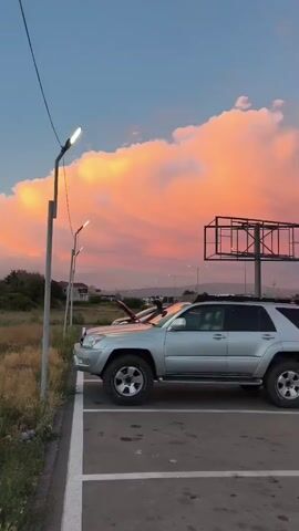 A silver SUV is parked in a parking lot. The sky is filled with vibrant pink and orange clouds during sunset. A billboard is visible behind the SUV. The road in front of the parking lot has traffic.