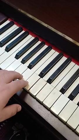 A close-up, slightly angled, shot focuses on the right hand of an individual playing a portion of a piano keyboard. The keyboard has white and black keys, and some of the white keys appear to be discolored. The hand is light-skinned and the fingernails are trimmed. The hand moves across the keys, playing a sequence of notes. The piano has a dark brown wooden casing with a red-colored area visible underneath the keys. The lighting in the video appears to be ambient.