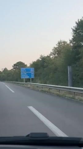 The video shows a view from a vehicle driving on a highway in France. A blue signpost with white text indicates distances to cities: Lyon (383 km), Paris (288 km), Dijon (191 km), Troyes (163 km), and Chaumont (126 km). Trees and foliage line the right side of the highway. Traffic is visible ahead, including trucks and cars. The sky is a light, hazy color, suggesting it is either early morning or late evening. The highway has clear lane markings.