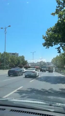 The video shows a view from inside a car driving on a multi-lane road under a clear blue sky. Several cars and a truck are visible on the road ahead. Trees line the right side of the road, and buildings can be seen in the distance. The car's dashboard is visible in the foreground.
