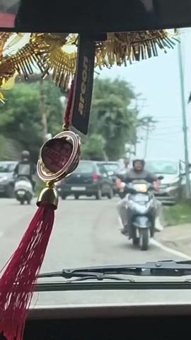 A first-person perspective shot from inside a vehicle looking out the windshield at a street scene. A red tassel and a gold ornament hang from the rearview mirror, partially obscuring the view. The road is busy with traffic, including cars, scooters, and a truck. Trees and power lines are visible along the side of the road. The lighting suggests it is daytime. The vehicle appears to be moving.