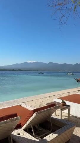 The video shows a serene beach scene with turquoise waters and distant mountains under a clear blue sky. In the foreground, lounge chairs with orange cushions are set up on a paved area bordering the sand. A small table with a potted plant sits next to one of the chairs. The calm ocean features a few scattered swimmers and a motorboat cruising across the water. The lush green coastline of the distant landmass is visible, with a hazy mountain range dominating the horizon, capped by a wispy cloud. The overall impression is one of a peaceful tropical vacation destination.
