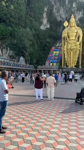 The video captures a wide shot of the entrance to the Batu Caves in Gombak, Selangor, Malaysia. In the foreground, a tiled plaza with a distinctive red, black, and white/beige 3D cube pattern is visible, where several people are standing and walking.

The most prominent feature is the colossal 42.7-meter-tall golden statue of Lord Murugan, standing majestically on the right side of the frame. To its left, a long, steep staircase with 272 steps leads up to the cave entrance. These steps are strikingly painted in various bright colors, including red, blue, green, and yellow, with people ascending and descending them.

In the mid-ground, at the base of the stairs, there's a bustling area with many visitors and some smaller, intricately decorated Hindu temple structures. Pigeons are also seen gathered on the ground in this area. A lush, green mountain or cliff face covered in trees forms the backdrop, enveloping the cave entrance and the upper parts of the steps.

In the immediate foreground, to the left, a man in a light blue shirt and jeans is standing with his hands pressed together, possibly in prayer or reverence, while holding a red bag. Another man and woman are standing in the center, facing the steps and the statue. On the far right, a baby stroller is parked.

The scene is brightly lit by natural daylight, indicating a clear, sunny day. The overall atmosphere appears busy with a mix of devotees and tourists visiting the iconic religious site.