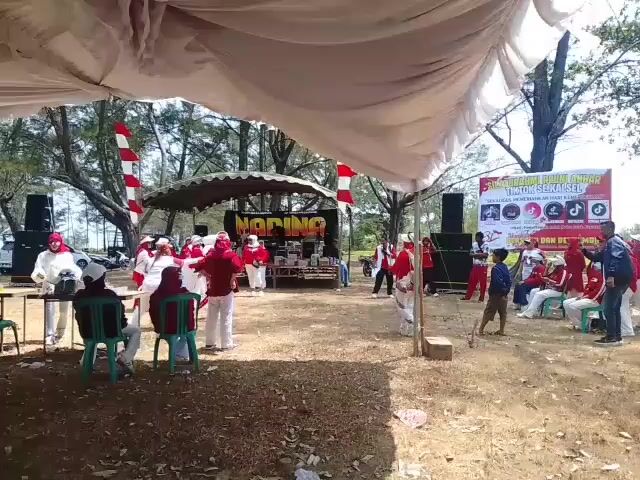 The video shows an outdoor event, possibly a performance or gathering, under a large white tent. In the foreground, a few people are seated on chairs, looking towards a central area. The central area features a stage or platform with "NADINO" written on it in large letters. A group of people, mostly dressed in red and white, are gathered around the stage, some appearing to be performing or preparing to perform. There are several speakers and what appear to be sound equipment. A banner is visible in the background with text and images, including social media logos. The environment is outdoors with trees and dry ground, suggesting a park or open field. The lighting indicates it is daytime.